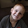 child, boy, smiling, portrait, indoors, couch, seated, spiderman, superhero, t_shirt, clothing, brown_hair, short_hair, fair_skin, head_tilt, happy, camera_facing, home, hanging_clothes, casual