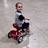 child, toddler, tricycle, riding, smiling, portrait, indoor, polished_floor, hall, adults, table, chair, traffic_cone, casual_clothing, shorts, shoes, play, vehicle, reflection, architecture