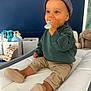 Charles participe au concours pour gagner de l'argent avec cette photo : baby, child, teether, cap, green_sweater, beige_pants, indoor, changing_pad, nursery, plush_toy, blue_wall, baby_care_products, footwear, sitting, cute, infant, smiling, person, headwear, portrait