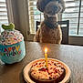 dog, birthday_cake, candle, plush_toy, happy_birthday, table, chair, window, blinds, indoor, celebration, pet, curly_fur, food, plate, light, decor, animal, birthday, waiting