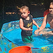 Jamal is registered to the contest to win money with this photo: child, pool, water, outdoor, summer, sunlight, orange_bucket, greens, playing, splash, tank_top, shorts, nature, fun, kids, sunny_day, backyard, water_toy, happy, sibling