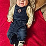 baby, smiling, denim_overalls, sneakers, red_blanket, cushion, infant, happy, lying_down, cute, child, portrait, indoor, clothing, footwear, soft_texture, young, person, casual_wear, comfortable
