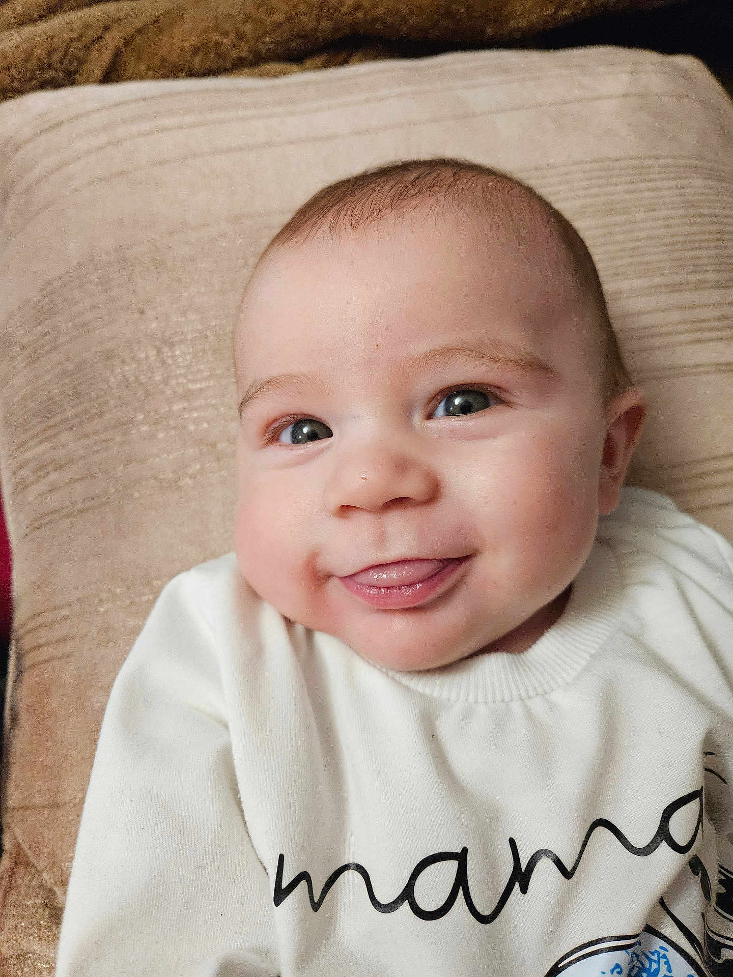 Renato participe au concours pour gagner de l'argent avec cette photo : baby, infant, face, smile, tongue_out, white_shirt, text_on_clothing, cushion, beige, skin, head, eyes, cheeks, closeup, happy, child, portrait, indoors, person, clothing