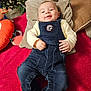 baby, denim_overalls, cushion, red_blanket, smiling, indoor, baby_clothing, child, face, happy, shoes, toy, christmas_tree, home_decor, pillow, person, infant, cute, resting, cozy