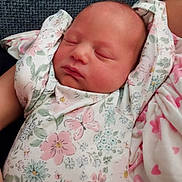 Saige is registered to the contest to win money with this photo: baby, newborn, sleeping, floral_onesie, infant, arms_up, blanket, pink_hearts, cozy, peaceful, face, head, skin, person, portrait, resting, soft_texture, blue_background, closeup, indoors