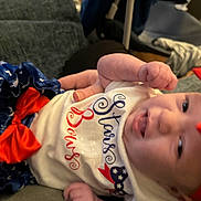Saige joined the competition — help win amazing prizes! baby, blue_skirt, celebration, child, clothing, couch, cute, face, festive, hand, happy, indoor, infant, person, portrait, red_bow, smile, stars, tongue, white_shirt