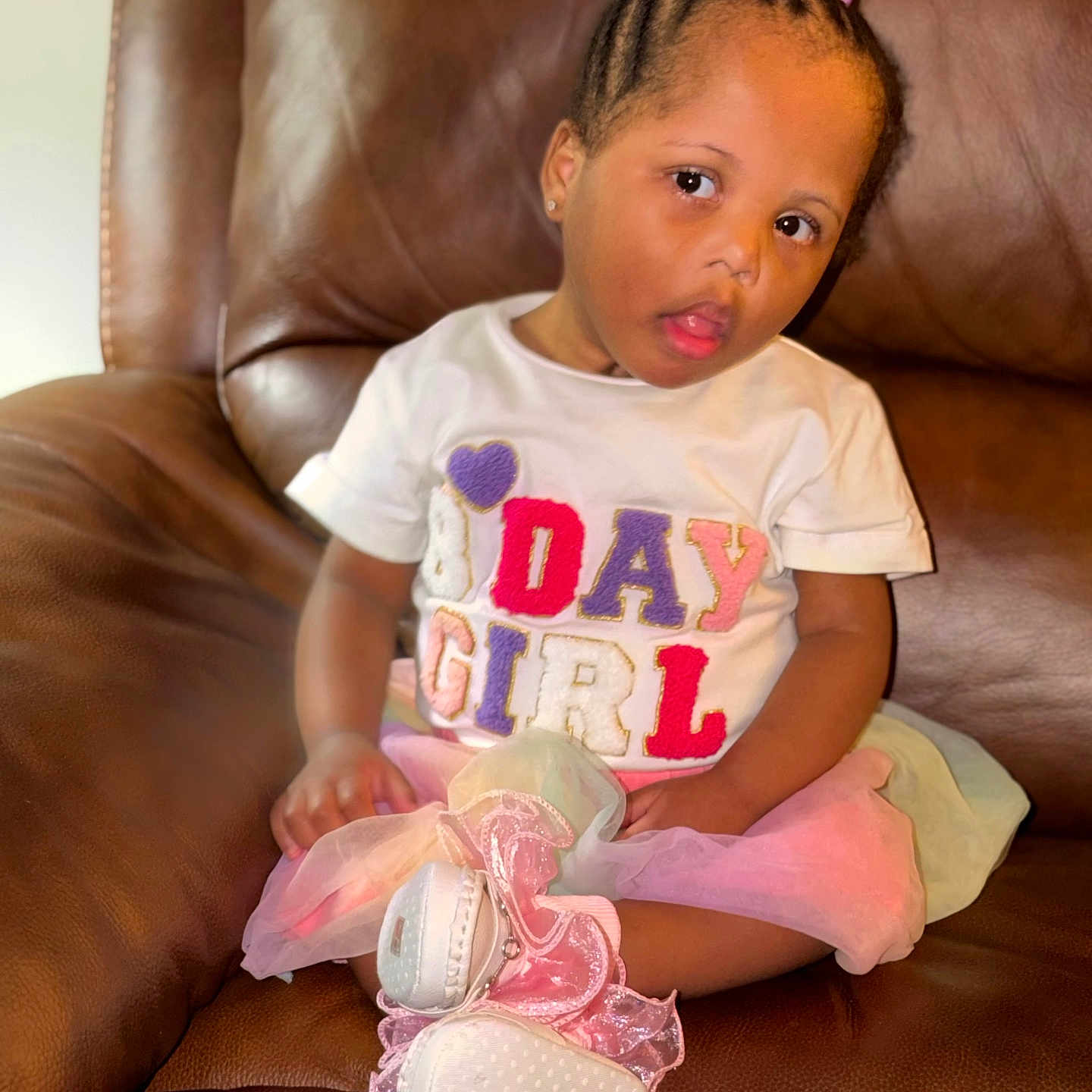 Elyse is registered to the contest to win money with this photo: child, girl, toddler, portrait, sitting, leather_couch, indoors, braids, hair_bows, earrings, white_shirt, colorful_clothing, skirt, sparkly_shoes, pink_shoes, fashion, serious_expression, face, furniture, home