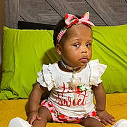 Elyse is registered to the contest to win money with this photo: baby, child, infant, headband, pink, strawberry, shirt, clothing, sitting, yellow_quilt, pillow, green_pillow, white_shoes, face, expression, indoor, cute, portrait, young_child, accessory