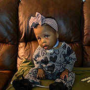 Elyse joined the competition — help win amazing prizes! toddler, child, headband, bow, heart_pattern, gray_clothing, sitting, leather_couch, green_blanket, black_shoes, earrings, indoor, curious_expression, person, baby, face, small_hands, couch_cushion, medical_tube, portrait