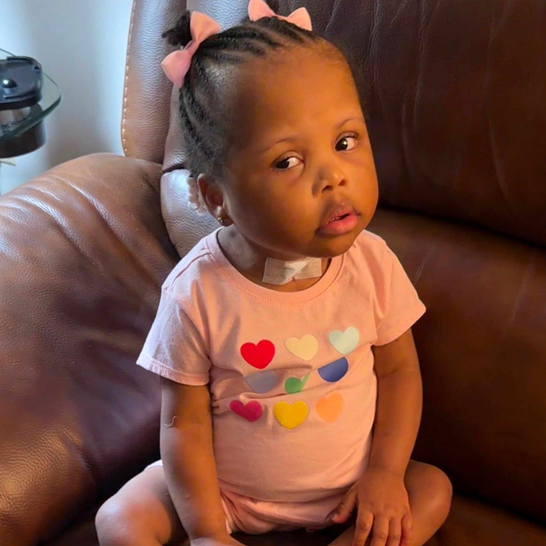 Elyse is registered to the contest to win money with this photo: toddler, child, braided_hair, pink_bows, pink_outfit, hearts, sitting, leather_chair, indoor, curious_expression, bandage, young_child, portrait, casual_clothing, cute, skin, face, hands, legs, home