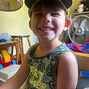Beau joined the competition — help win amazing prizes! child, boy, smiling, cap, tank_top, teeth, indoor, fan, toy, wooden_furniture, mirror, yellow, green, happy, portrait, casual_clothing, playful, room, furniture, person
