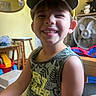 child, boy, smiling, cap, tank_top, teeth, indoor, fan, toy, wooden_furniture, mirror, yellow, green, happy, portrait, casual_clothing, playful, room, furniture, person