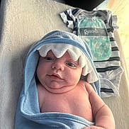 Ryder is registered to the contest to win money with this photo: baby, infant, blue_towel, hooded_towel, shark_design, onesie, striped_clothing, text_on_clothing, soft_blanket, cream_blanket, cute, chubby_cheeks, lying_down, indoors, close_up, child, newborn, smiling, portrait, casual