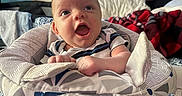 Ryder is registered to the contest to win money with this photo: air_conditioner, baby, baby_seat, blanket, child, clothing, colorful, expression, face, furniture, happy, head, home, indoor, infant, painting, patterned_fabric, person, smiling, window