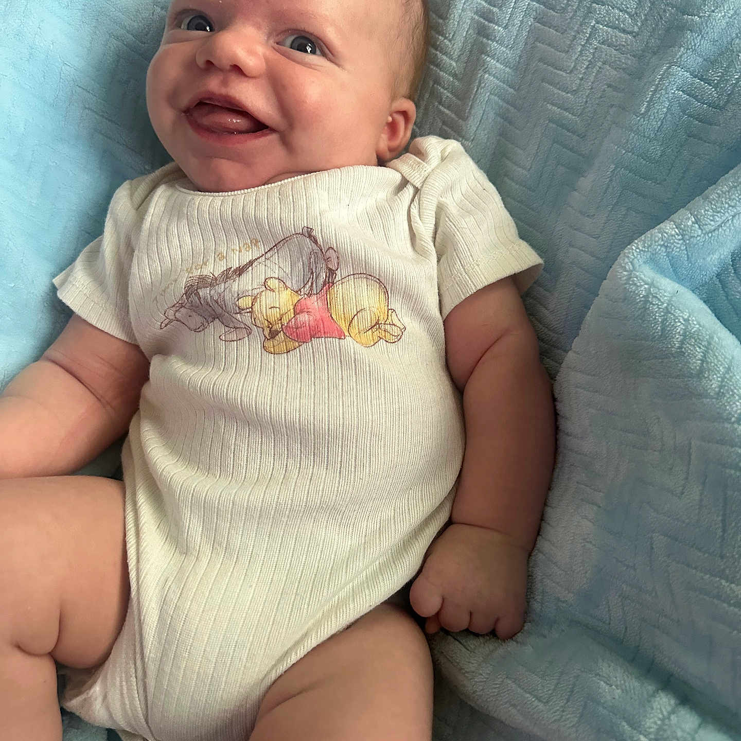 Ryder is registered to the contest to win money with this photo: baby, infant, smile, onesie, winnie_the_pooh, blanket, blue, happy, cute, child, portrait, skin, face, arm, leg, clothing, lying_down, soft_texture, infant_clothing, toddler