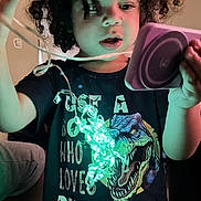 Jonah is registered to the contest to win money with this photo: child, curly_hair, tshirt, dinosaur, lights, string_lights, glowing, indoor, purple_device, curious, portrait, young, person, black_tshirt, room, wall, couch, hand, expression, dinosaur_graphic