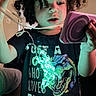 child, curly_hair, tshirt, dinosaur, lights, string_lights, glowing, indoor, purple_device, curious, portrait, young, person, black_tshirt, room, wall, couch, hand, expression, dinosaur_graphic