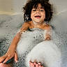 child, bath, foam, bubbles, curly_hair, smiling, happy, playful, water, soap, tiled_wall, foot, hand, skin, bathtub, bubble_bath, indoor, person, childhood, cleaning