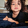 child, toddler, curly_hair, black_shirt, blanket, indoor, face, portrait, expression, hands, young, person, candid, skin, bedroom, relaxed, resting, casual, cute, curly