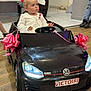 toddler, child, toy_car, volkswagen, indoor, pink_ribbon, floor, curious, white_jacket, steering_wheel, head, face, person, vehicle, small_car, flooring, event, decor, young_child, casual