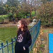 Sirine a rejoint le concours — aidez-le/la à gagner de superbes lots ! child, girl, curly_hair, black_hoodie, pink_leggings, pond, waterfall, blue_fence, trees, nature, outdoor, smile, side_view, casual, park, grass, rocks, water, overcast, bracelet