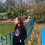 child, girl, curly_hair, black_hoodie, pink_leggings, pond, waterfall, blue_fence, trees, nature, outdoor, smile, side_view, casual, park, grass, rocks, water, overcast, bracelet