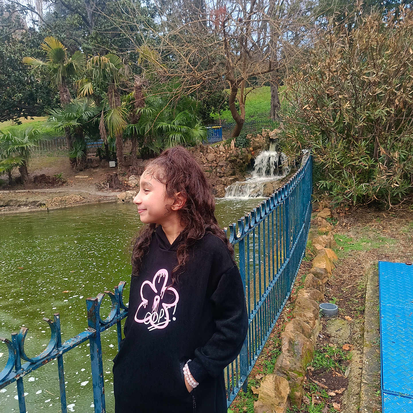 Sirine a rejoint le concours — aidez-le/la à gagner de superbes lots ! black_hoodie, blue_fence, bracelet, casual, child, curly_hair, girl, grass, nature, outdoor, overcast, park, pink_leggings, pond, rocks, side_view, smile, trees, water, waterfall