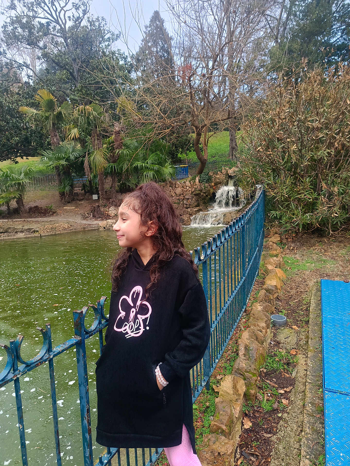 Sirine a rejoint le concours — aidez-le/la à gagner de superbes lots ! child, girl, curly_hair, black_hoodie, pink_leggings, pond, waterfall, blue_fence, trees, nature, outdoor, smile, side_view, casual, park, grass, rocks, water, overcast, bracelet