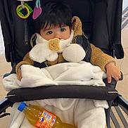 Kent participe au concours pour gagner de l'argent avec cette photo : baby, black_hair, blanket, bottle, brown_clothing, child, cozy, curious, cute, face, fanta, hands, indoor, orange_soda, plush_toy, seated, soft_toy, stroller, toddler, warm_clothing