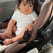 Matías Felix is registered to the contest to win money with this photo: baby, child, car_seat, car_interior, seatbelt, white_shirt, curly_hair, cute, infant, sitting, looking, grip, buckle, window, vehicle, seat, toddler, indoors, expression, young