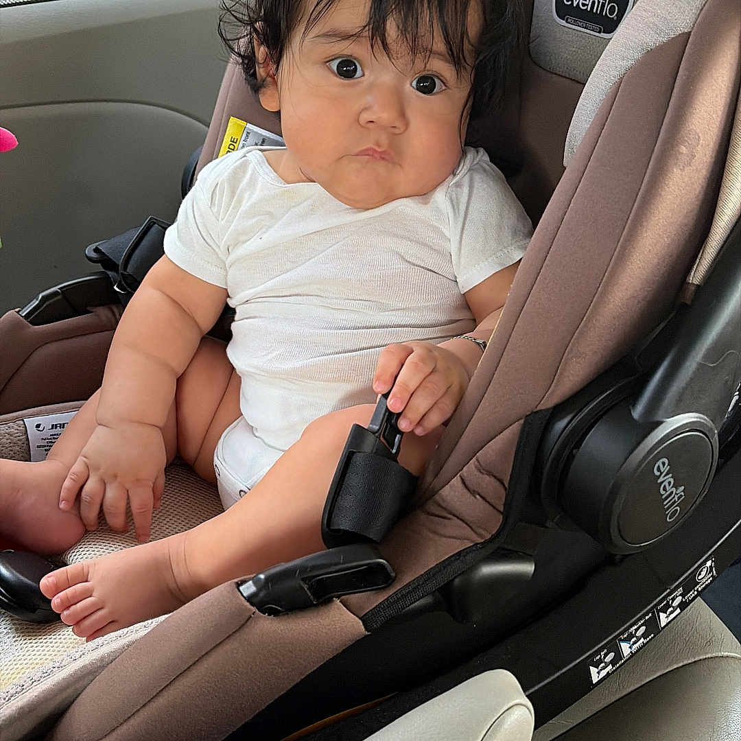 Matías Felix is registered to the contest to win money with this photo: baby, child, car_seat, car_interior, seatbelt, white_shirt, curly_hair, cute, infant, sitting, looking, grip, buckle, window, vehicle, seat, toddler, indoors, expression, young