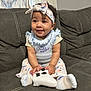 baby, child, girl, couch, controller, headband, smile, sitting, sock, clothing, curly_hair, indoor, person, cute, happy, game_controller, fabric, pattern, home, light
