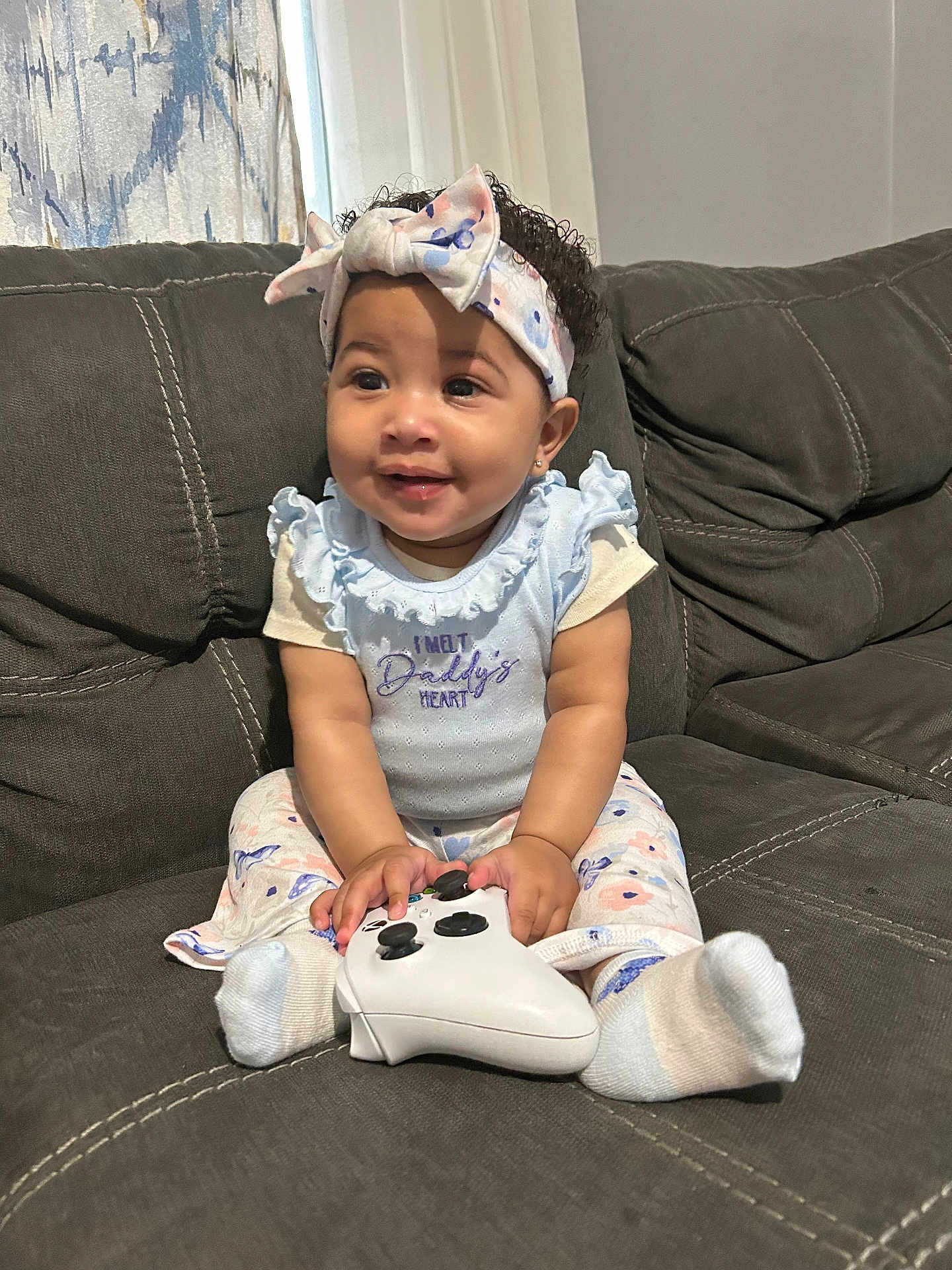 Journeylynn is registered to the contest to win money with this photo: baby, child, girl, couch, controller, headband, smile, sitting, sock, clothing, curly_hair, indoor, person, cute, happy, game_controller, fabric, pattern, home, light