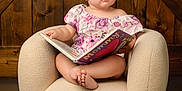 Maisie is registered to the contest to win money with this photo: baby, child, girl, bow, headband, book, reading, armchair, furniture, wooden_background, indoors, cute, curious, portrait, infant, pink, purple, floral_dress, barefoot, sitting