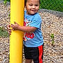 child, toddler, playing, yellow_pole, blue_shirt, shorts, sneakers, outdoor, playground, wood_chips, fence, grass, summer, happy, cute, smiling, person, casual_clothing, daytime, fun