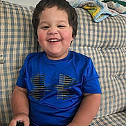 Grayson is registered to the contest to win money with this photo: blanket, blue_shirt, casual_clothing, child, chubby_cheeks, couch, hand, happy, holding_shoe, indoor, living_room, plaid_couch, portrait, seated, short_hair, smiling, sneaker, teeth, toddler, under_armour_logo