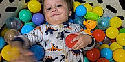 Grayson is registered to the contest to win money with this photo: ball_pit, blue_ball, child, colorful, cozy, dinosaur_pajamas, green_ball, happy, indoor, padded_enclosure, person, plastic_balls, play, purple_ball, red_ball, smile, toddler, toy, yellow_ball, young_child