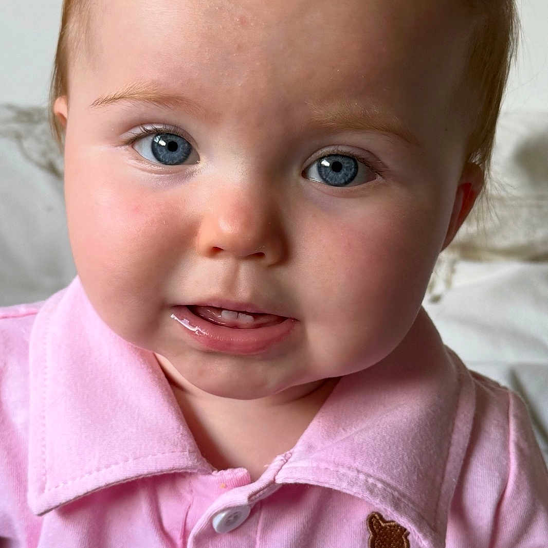 Isla is registered to the contest to win money with this photo: baby, blue_eyes, buttoned_shirt, child, close_up, collared_shirt, curious_expression, cute, face, head, indoor, infant, person, pink_shirt, portrait, skin, smile, soft_light, teddy_bear_patch, young_child