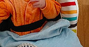 Nolan participe au concours pour gagner de l'argent avec cette photo : baby, blanket, blue_blanket, bouncer_seat, camera_watermark, child, closeup, curious_expression, eyes, fabric, face, hand, indoor, infant, onesie, orange_clothing, portrait, relaxed, striped_cushion, wooden_floor