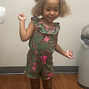 Azylah is registered to the contest to win money with this photo: casual, child, curly_hair, cute, floral_pattern, girl, green_clothing, happy, human, indoor, person, playful, romper, shorts, skin, smiling, standing, trash_can, wooden_floor, young