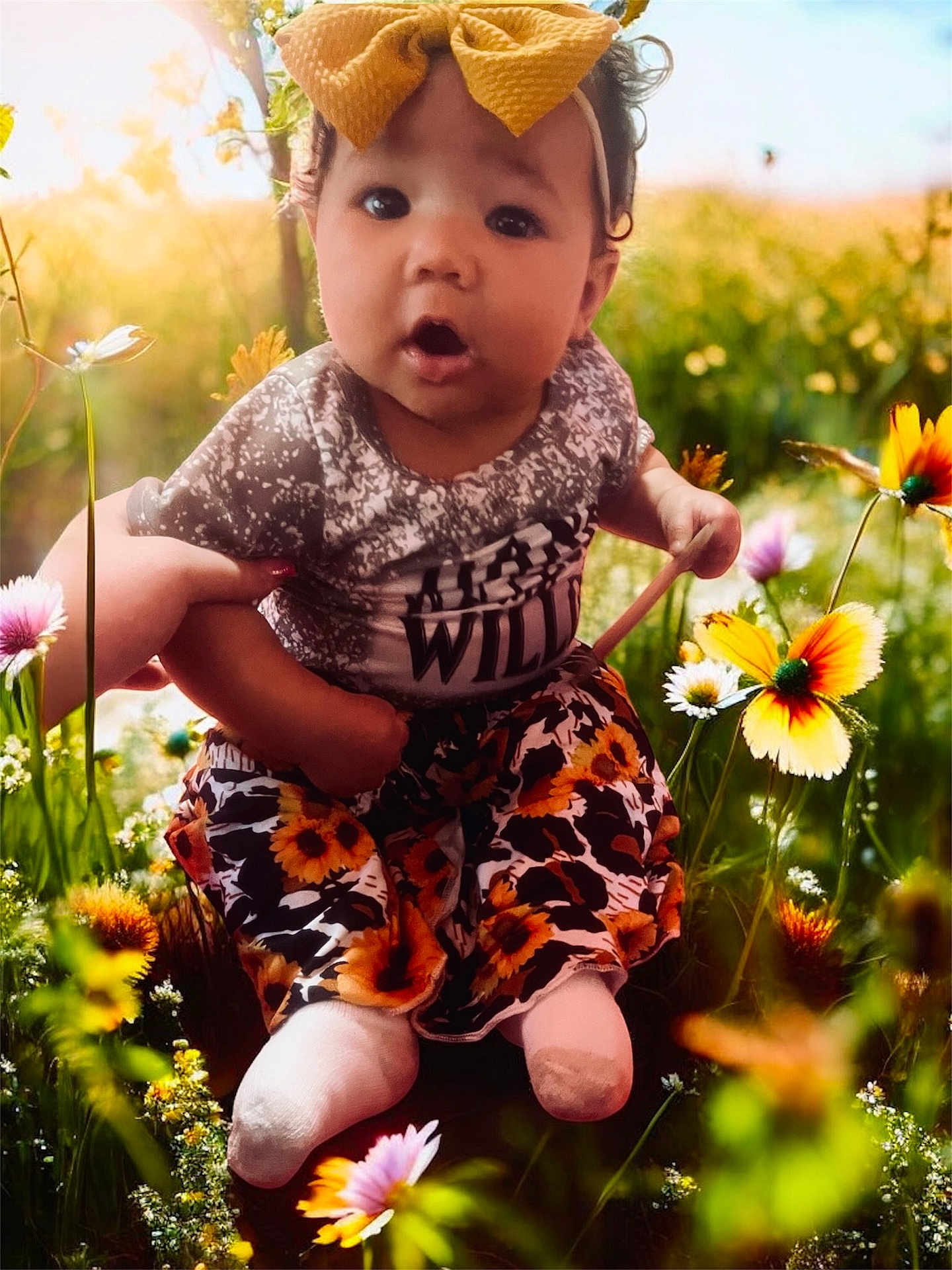 Oaklynn is registered to the contest to win money with this photo: baby, child, flower_field, yellow_bow, sunflower_pants, shirt, hand_holding, outdoor, nature, greenery, grass, colorful_flowers, sunlight, cute, portrait, infant, sitting, happy, spring, closeup