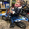 boots, child, container, curly_hair, floor, garage, girl, helmet, helmet_stand, indoor, jacket, jeans, motorbike, motorcycle, phone, scarf, shelf, smile, toy, young
