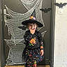 Luna participe au concours pour gagner de l'argent avec cette photo : bat, black_clothing, boots, child, costume, cute, decorations, door, festive, floor, halloween, hat, holiday, indoor, playful, pumpkin, smile, spider_web, tights, witch_costume