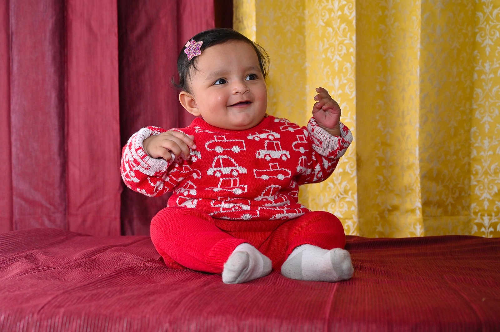 Utkarsha is registered to the contest to win money with this photo: baby, child, smiling, red_sweater, car_pattern, red_pants, white_socks, sitting, bedspread, maroon_curtain, yellow_curtain, indoor, happy, portrait, cute, infant, person, clothing, hairclip, decor