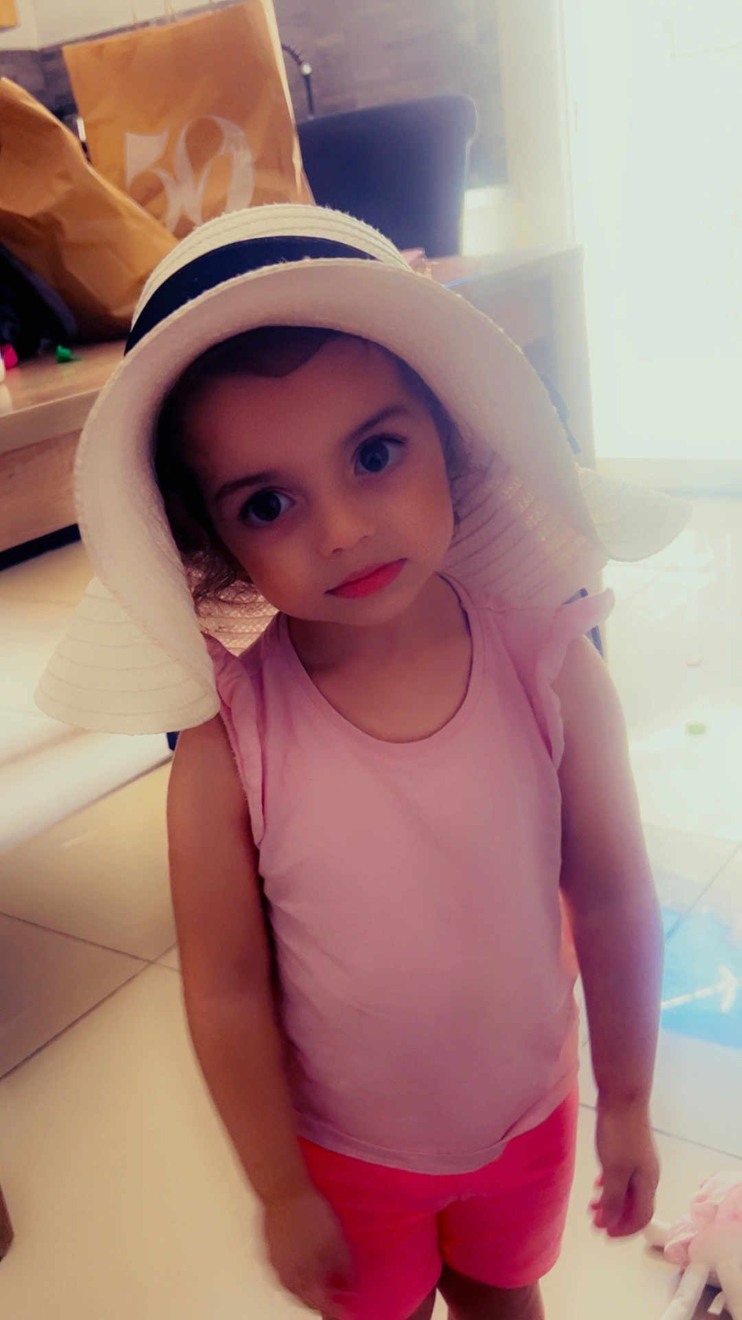 Layana participe au concours pour gagner de l'argent avec cette photo : child, girl, hat, pink_clothing, shorts, indoor, floor, expression, face, person, standing, curious, large_eyes, sunhat, casual, young, portrait, home, soft_lighting, toy