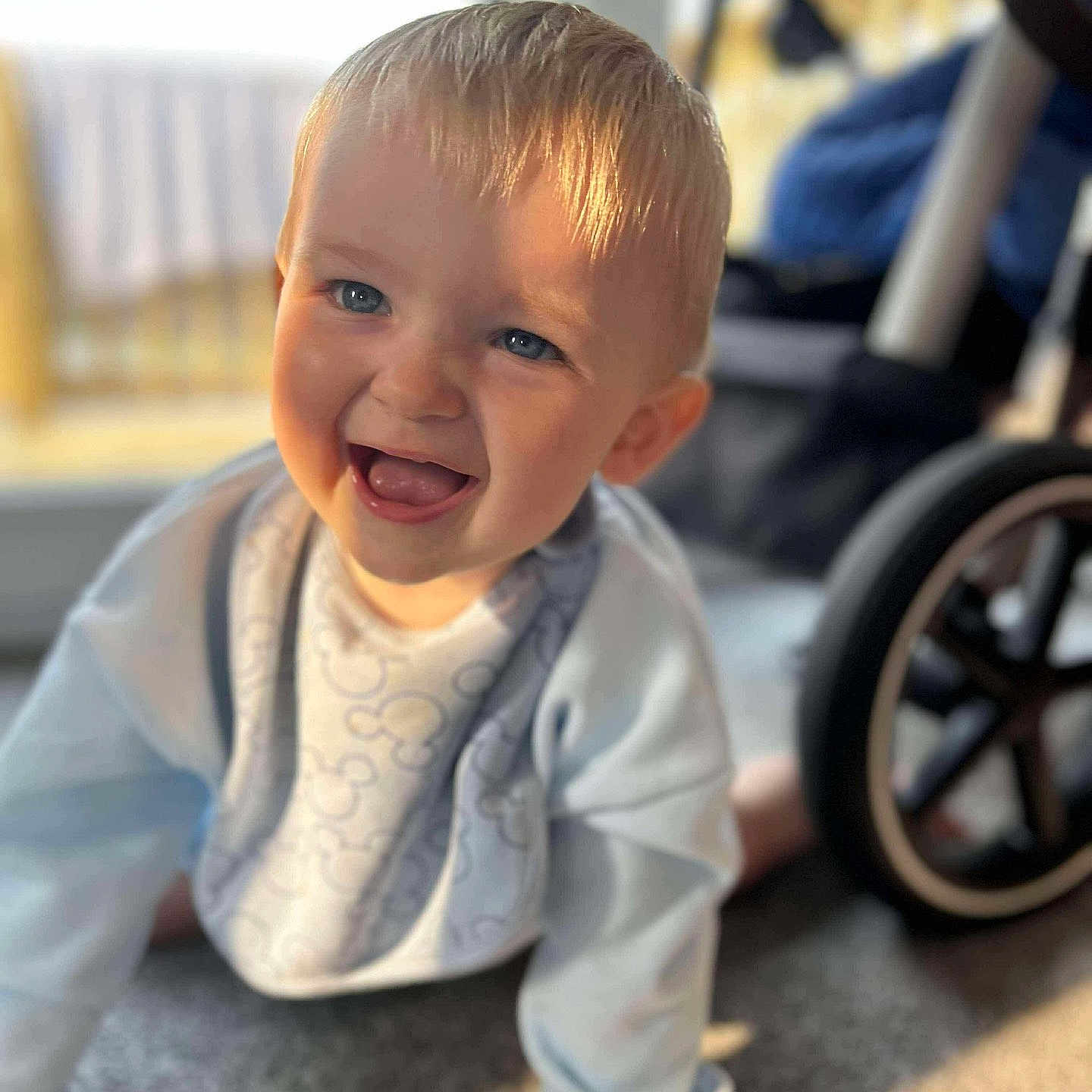 Lincoln is registered to the contest to win money with this photo: baby, bib, blonde_hair, blue_eyes, carpet, child, closeup, crawling, cute, happy, indoor, infant, light_blue_clothing, portrait, smiling, stroller, sunlight, toddler, wheel, young_child