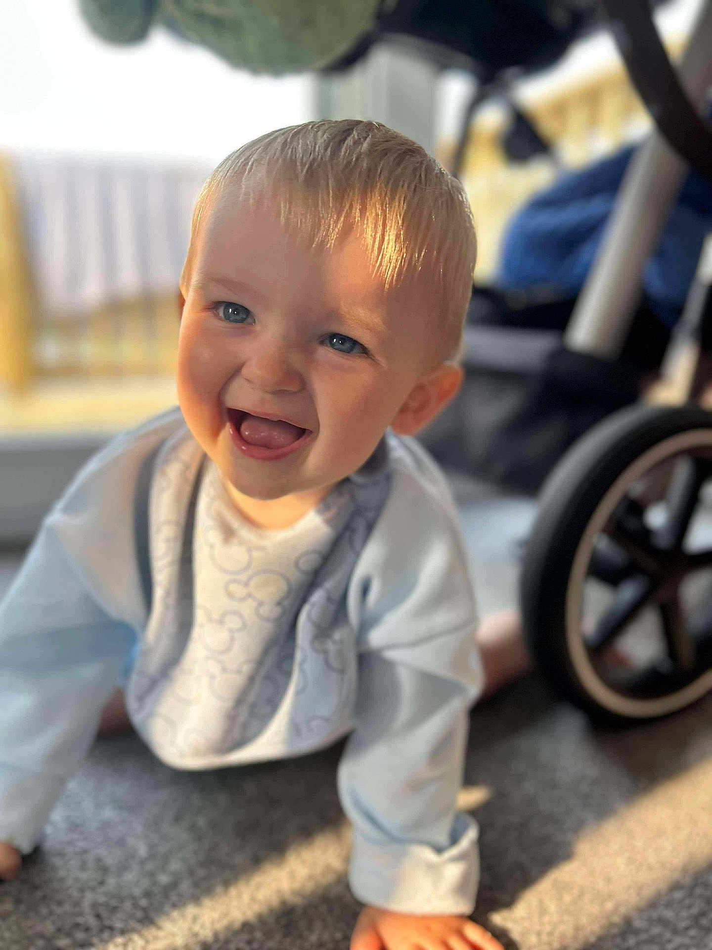 Lincoln is registered to the contest to win money with this photo: baby, child, crawling, smiling, blue_eyes, blonde_hair, bib, light_blue_clothing, carpet, indoor, sunlight, stroller, wheel, happy, cute, portrait, toddler, young_child, infant, closeup