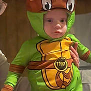 Maverick is registered to the contest to win money with this photo: baby, child, costume, teenage_mutant_ninja_turtles, green, hood, face, person, indoor, adult_hand, video_call, glasses, dark_background, bedroom, furniture, toy, cute, expressionless, party, holding
