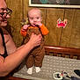 Maverick joined the competition — help win amazing prizes! baby, woman, glasses, brown_overalls, orange_shirt, changing_table, wood_paneling, stickers, sea_creatures, painting, hand, supporting, indoor, curious, playful, person, child, furniture, toy, socks