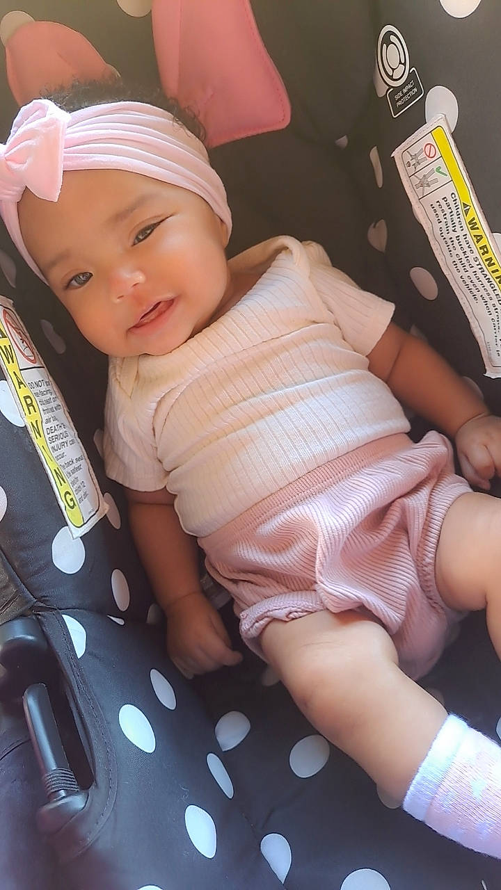 Aaliyah is registered to the contest to win money with this photo: baby, baby_toddler_clothing, cap, car_seat, cheek, child, comfort, happy, headwear, human_body, human_leg, lap, mouth, nail, pattern, person, pink, skin, smile, thigh
